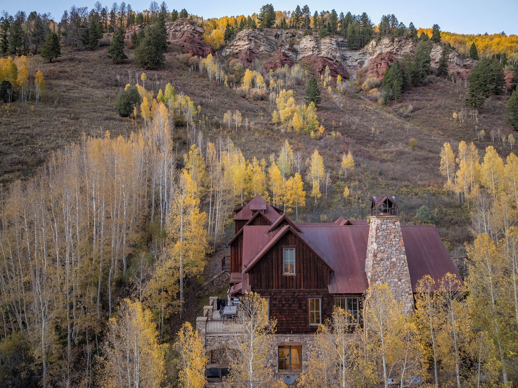 A rustic cabin with a stone chimney, surrounded by autumn-colored trees and a rocky hillside in the background.