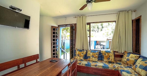A room with a wooden bench and a sofa with patterned cushions, a television mounted on the wall, and a ceiling fan. - Ground floor chalet 230 meters from Enseada Beach and 3 minutes from the sand. (UBATUBA)