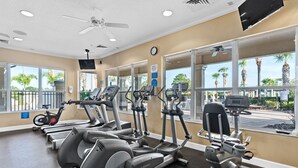 A gym with various exercise machines, a ceiling fan, and large windows with a view of palm trees. - RATED#1 BEST HOME Near Disney! LUXURY 5BEDROOM 5Bath VILLA ,GATED RESORT ,POOL (Kissimmee)