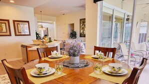 A dining area with a wooden table set for four, surrounded by chairs. There is a kitchen in the background with a white countertop and a sink. - RATED#1 BEST HOME Near Disney! LUXURY 5BEDROOM 5Bath VILLA ,GATED RESORT ,POOL (Kissimmee)