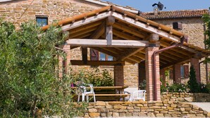 Ein Steinhaus mit einer Holzpergola, Außenbestuhlung und einer Steinsäule.