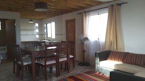 Un salón amplio con una mesa de comedor, sillas, un sofá, un colorido tapete a rayas, un ventilador de pie y una cocina al fondo.