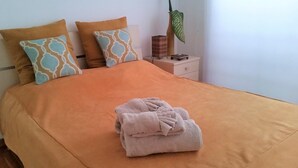 Una cama bien hecha con una colcha naranja y dos almohadas estampadas, una toalla doblada sobre la cama, una mesita de noche con una lámpara y una planta en maceta.