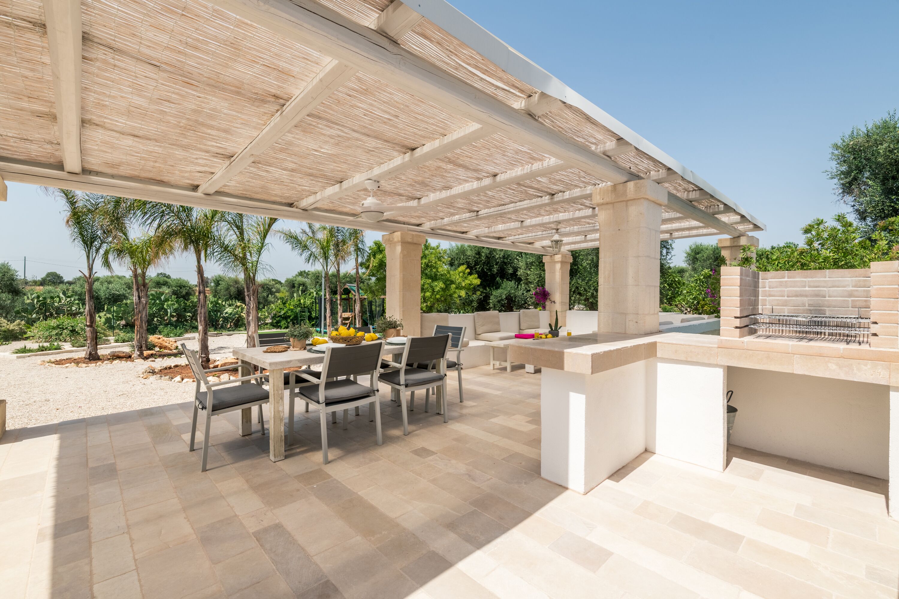 Patio esterno con pergola, tavolo e sedie da pranzo, barbecue e vista su palme e vegetazione.