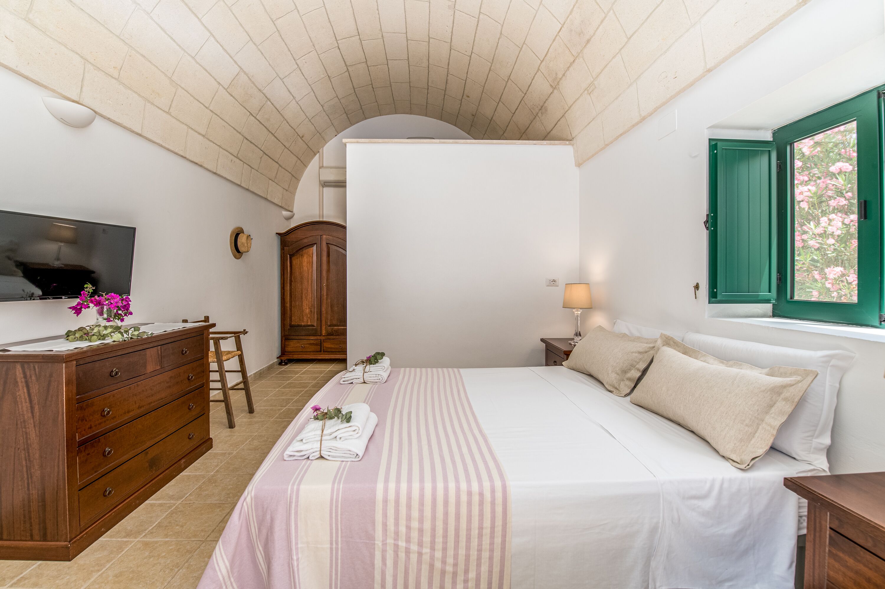 A bedroom with a bed, wooden dresser, TV, and a window with green shutters.