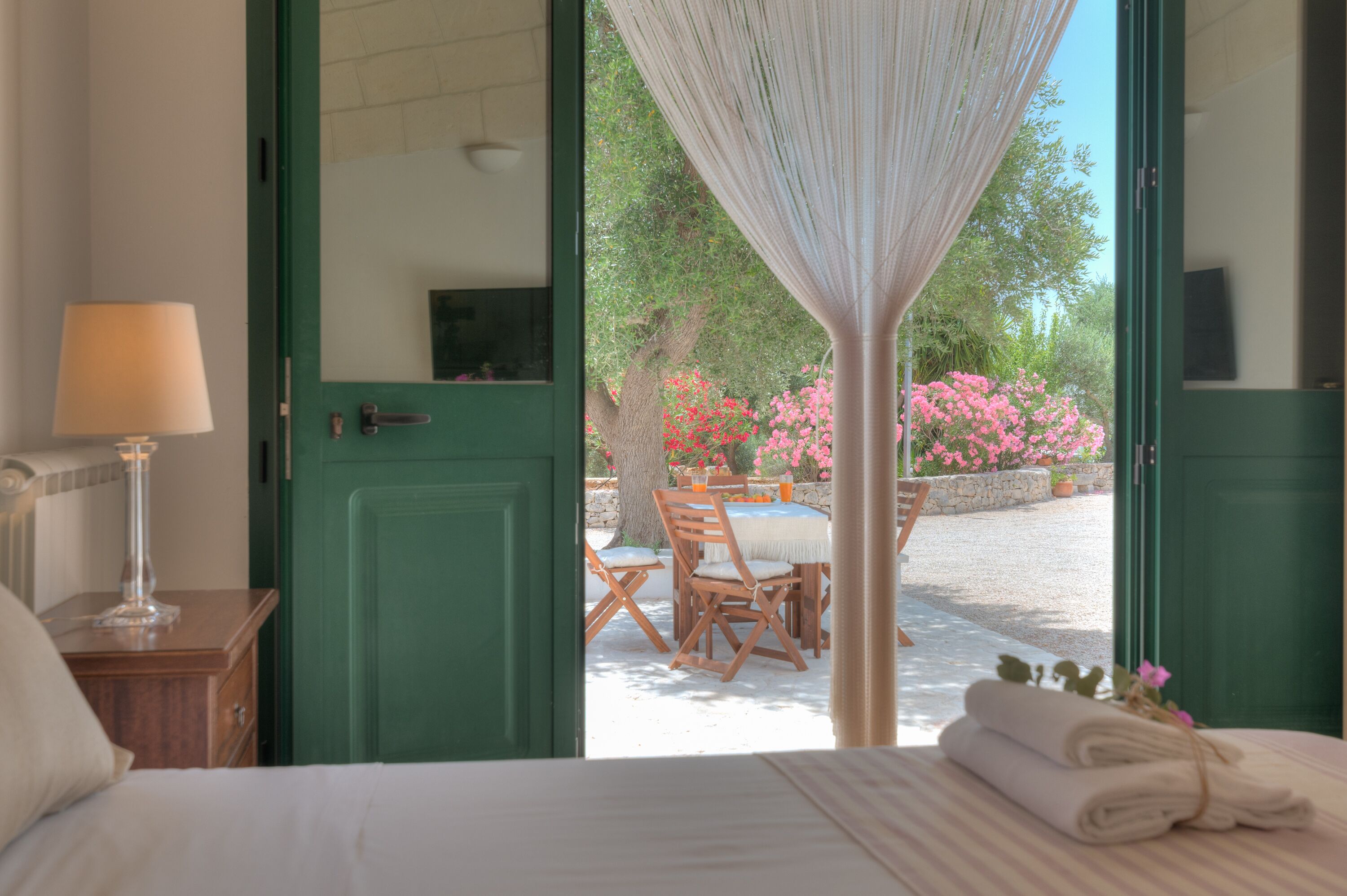 A bedroom with a bed, a nightstand with a lamp, and a view of an outdoor dining area with a table and chairs.