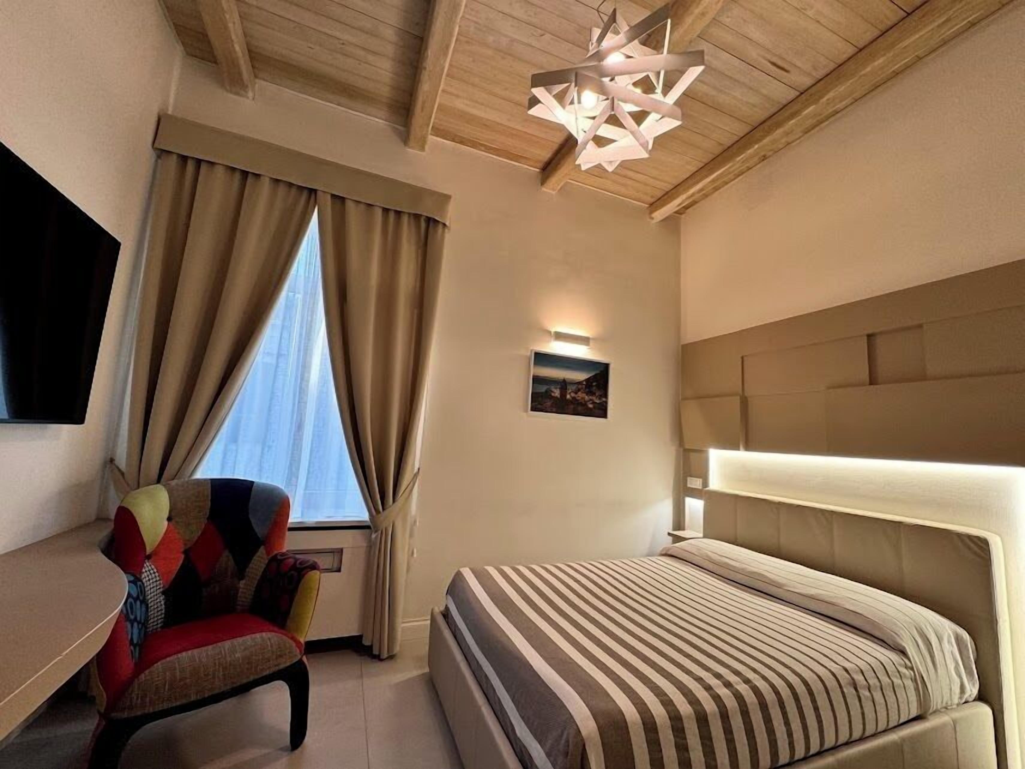 Een hotelkamer met een bed, een stoel, een televisie, een raam met gordijnen en een plafondlamp.