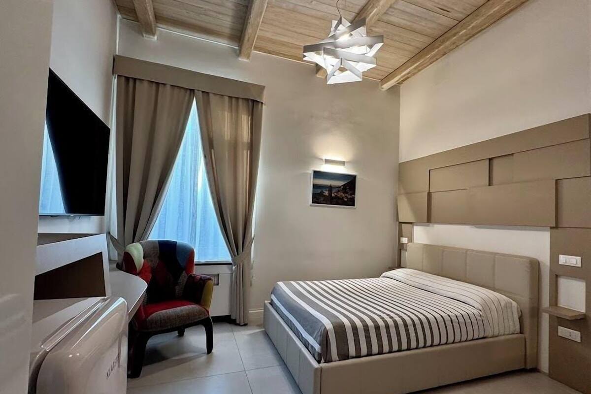 Moderni hotellihuone, jossa on suuri sänky, tuoli, litteänäyttöinen televisio ja ainutlaatuinen kattovalaisin.