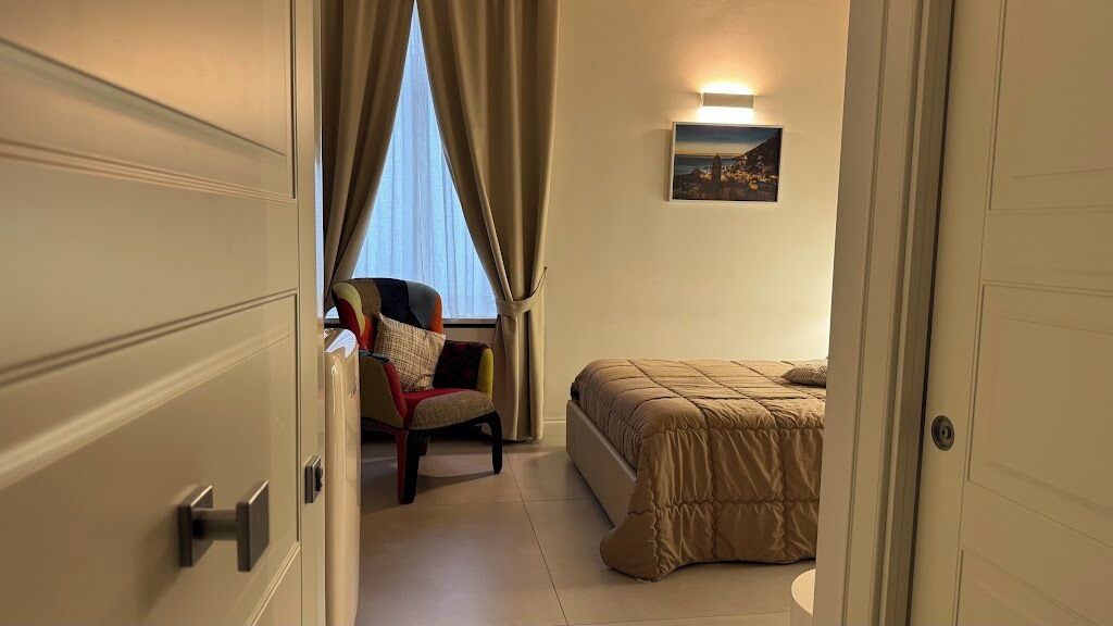 A hotel room with a bed, a chair, a window with curtains, and a wall-mounted light.