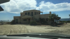 Un bâtiment à deux étages, doté d’un toit plat et d’une seule cheminée, entouré d’une zone sablonneuse où la végétation est clairsemée.