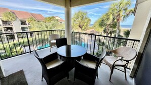 Un balcón con una mesa redonda y cuatro sillas de mimbre, con vista a una piscina y palmeras.
