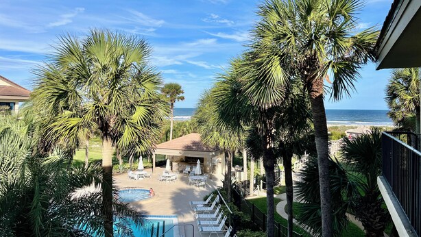 Zona de piscina con sillas de descanso y sombrillas, rodeada de palmeras y con vistas a una playa.