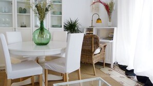 A dining area with a round table, white chairs, a green vase with flowers, a wicker chair, a lamp, and a bookshelf. - Very luminous apartment in the corner of the Cathedral, in the Arenal district (Sevilla)