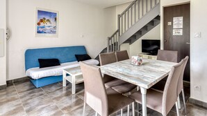 Una habitación con un sofá azul, una mesa de comedor con sillas, un televisor y una escalera.