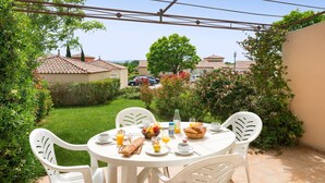Une table ronde entourée de chaises blanches, installée à l’extérieur, au milieu de la verdure, sous un ciel dégagé.