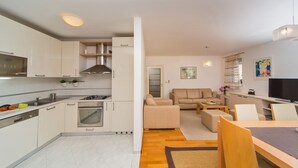 Une cuisine moderne avec des meubles blancs, une cuisinière et un évier. Le salon est équipé d’un canapé beige et d’un téléviseur.