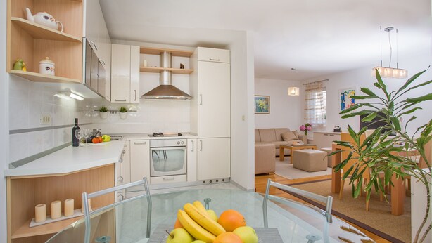 Une cuisine moderne avec des meubles blancs, une hotte en acier inoxydable et une table à manger en verre agrémentée d’un présentoir de fruits.