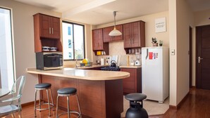A modern kitchen with wooden cabinets, a large island, bar stools, a refrigerator, and a window providing natural light. - Yellow Gem Apt - Campoverde (Cochabamba)