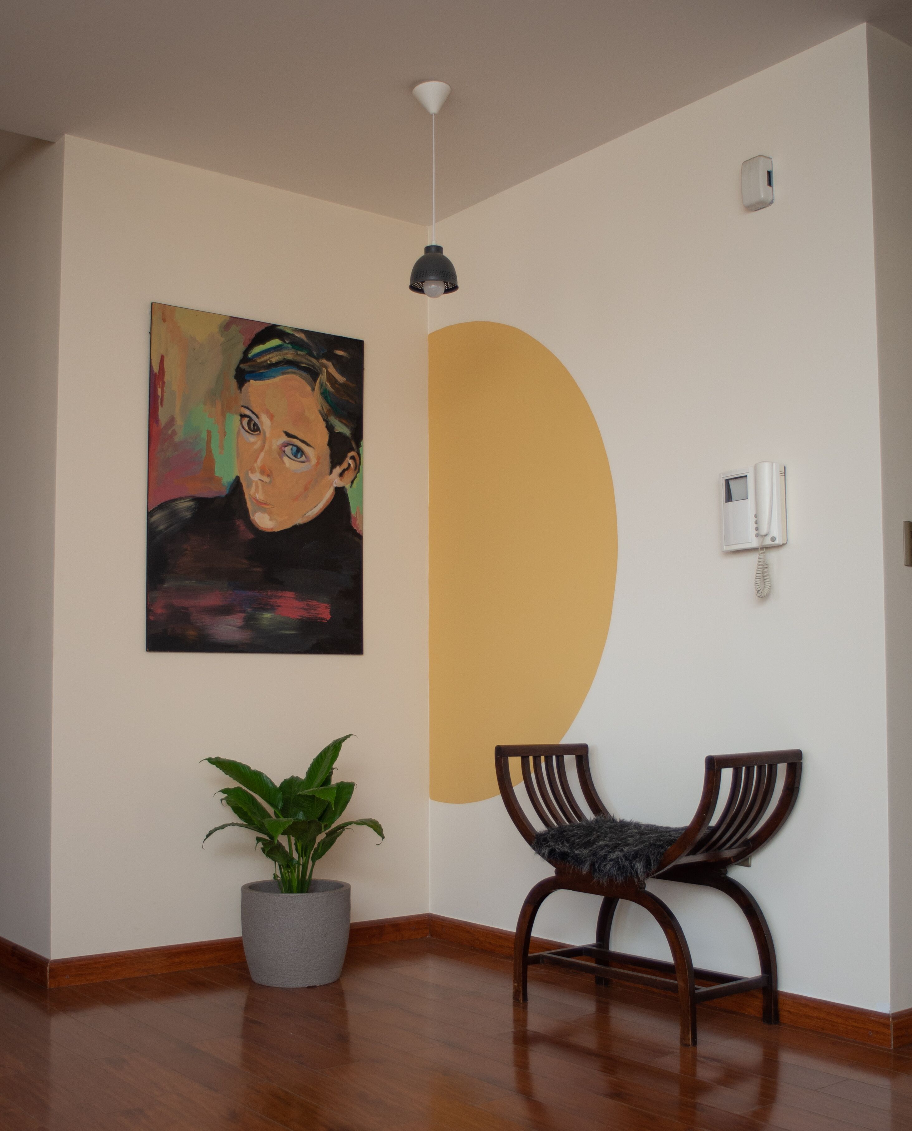 Una habitación con suelo de madera, una planta en maceta, una silla, un cuadro, un teléfono en la pared y una lámpara en el techo.