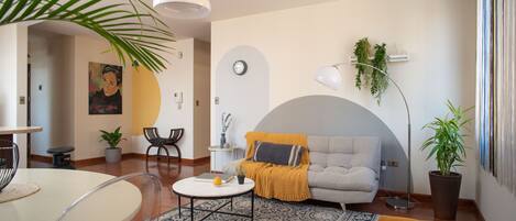 Un salon moderne avec un canapé, une table basse et des fauteuils. On y trouve également un coin repas avec une table et des chaises. La pièce est agrémentée d’une grande plante et d’une horloge murale.