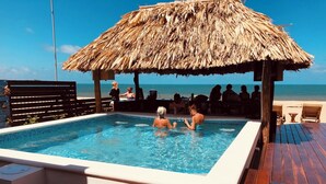 A pool area with a thatched roof structure, wooden deck, and a view of the ocean. - Economy Studio beachfront with swim up pool bar (Placencia)