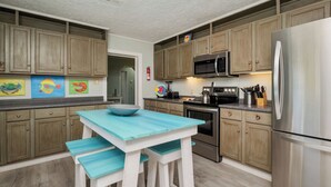 Een keuken met houten kastjes, een roestvrijstalen koelkast, een magnetron en een fornuis. Er staat een blauwe tafel met twee stoelen en een kom op.