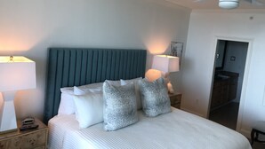 Quarto bem arrumado com uma cama grande, duas mesas de cabeceira com abajures, um ventilador de teto e uma porta que dá para outro cômodo.