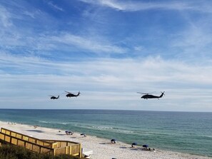 Una playa con un paseo marítimo de madera y gente tomando el sol. Tres helicópteros vuelan por encima.