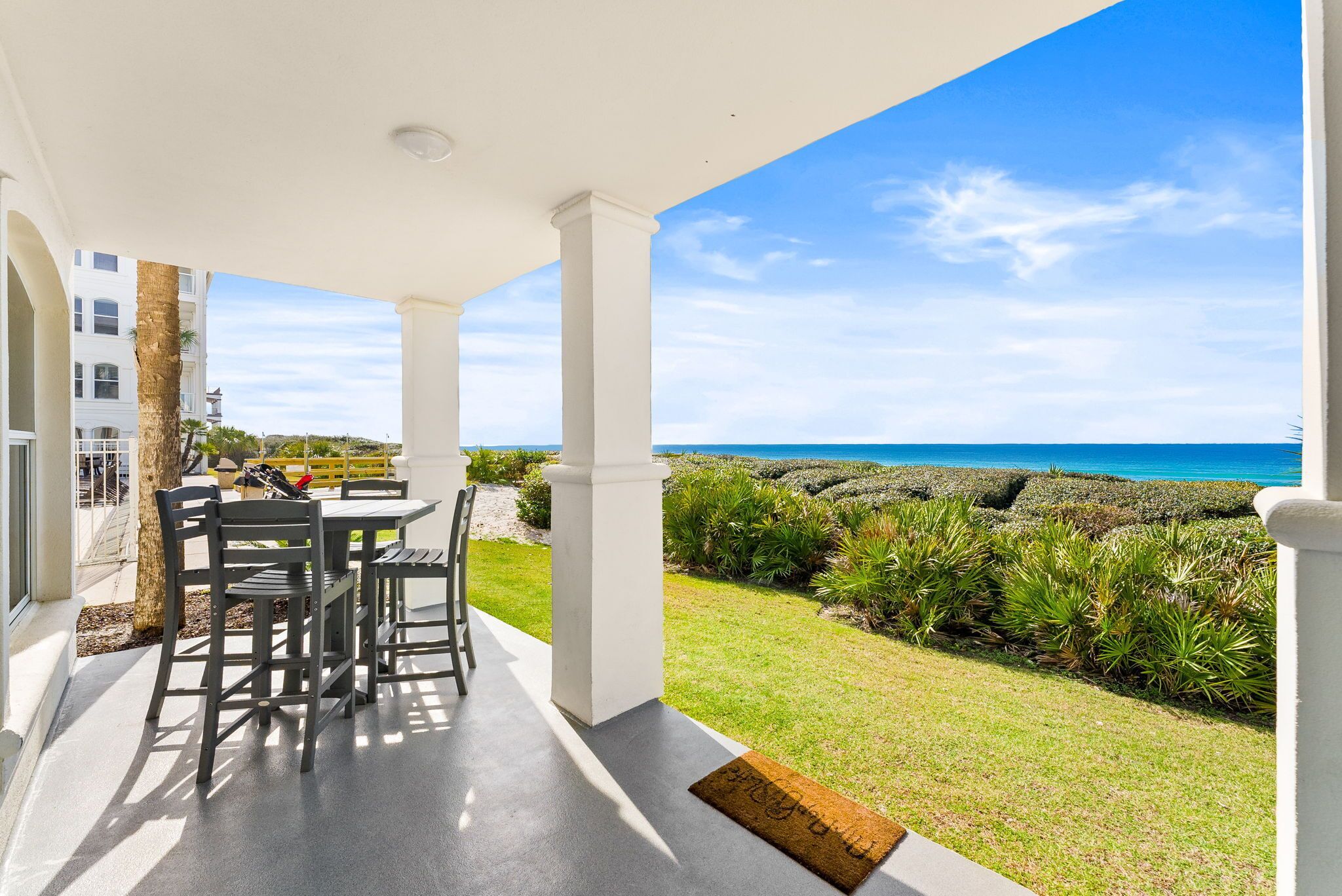 A covered patio with a view of the ocean, a white building, and a well-maintained lawn.