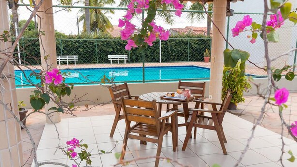 A patio with a pool, wooden chairs, and a table with a drink. - Maria Quitéria Hotel e Flat (Feira de Santana)