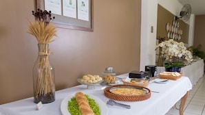A buffet table with various food items, a vase with dried plants, and a fan in the background. - Maria Quitéria Hotel e Flat (Feira de Santana)