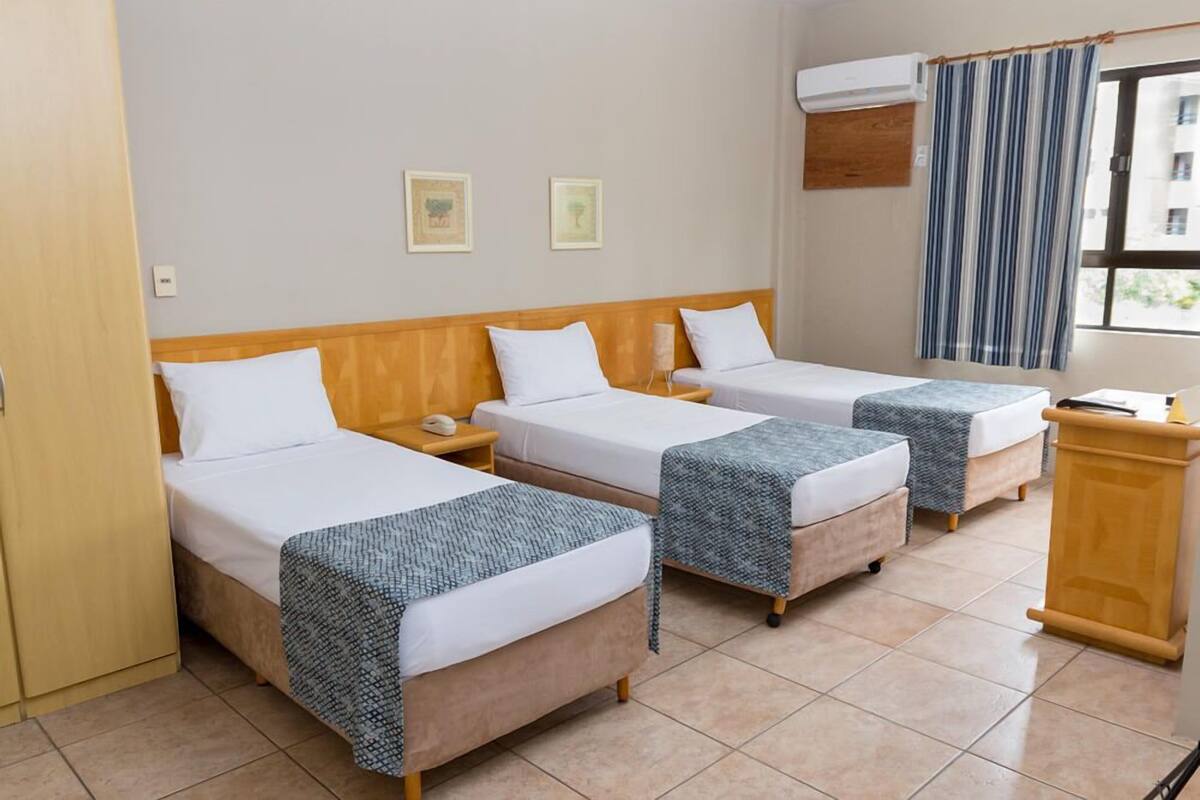 Une chambre d’hôtel avec trois lits simples, une tête de lit en bois, une table de chevet, un bureau et une fenêtre avec des rideaux.