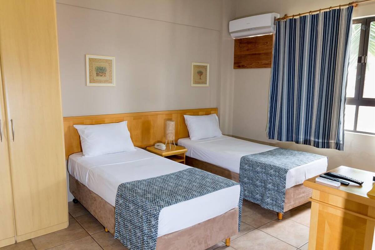 Une chambre d’hôtel avec deux lits, une armoire en bois, un bureau avec une chaise et une fenêtre avec un rideau à rayures.