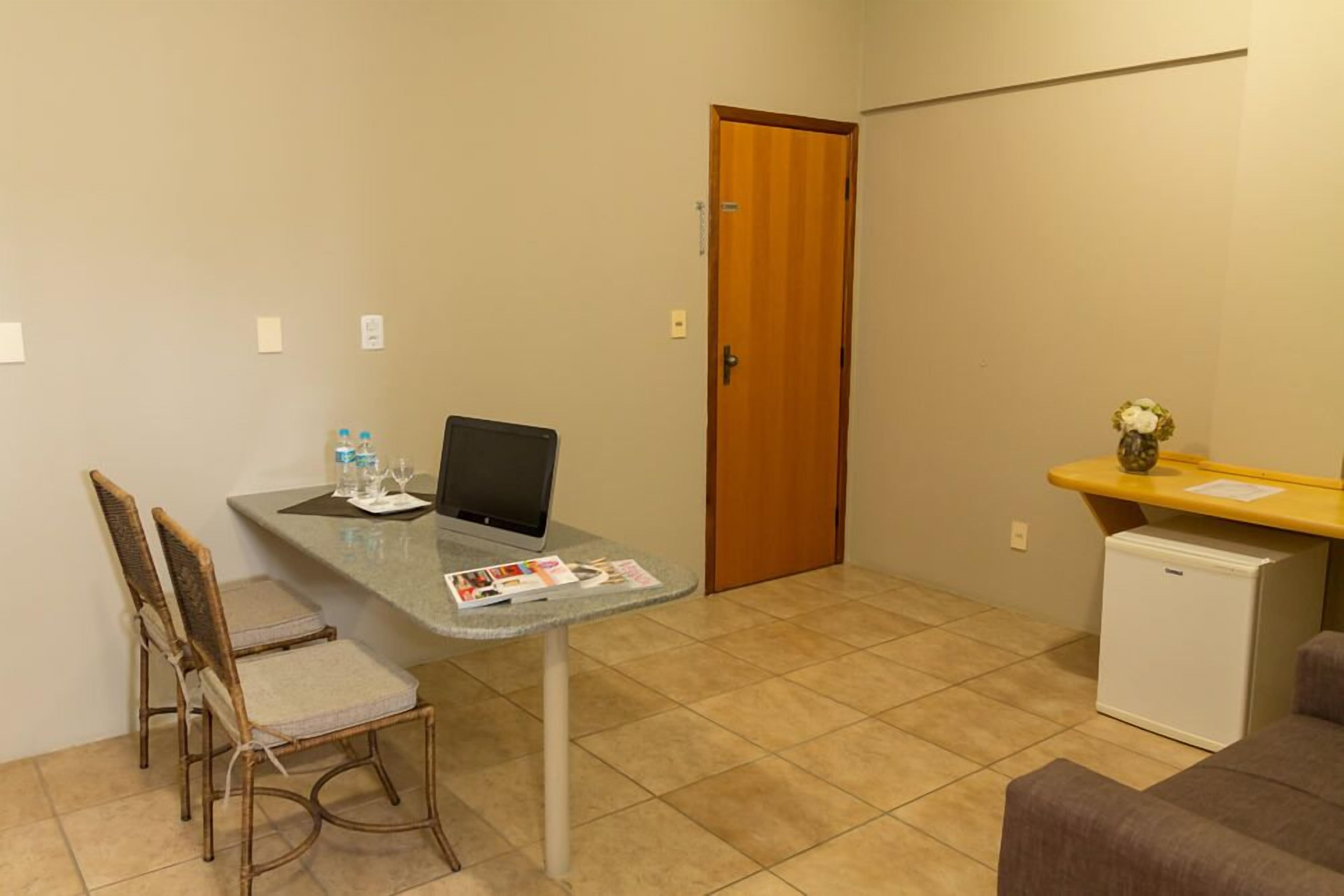 A small room with a kitchenette, a dining area with two chairs, a laptop on a table, and a refrigerator.