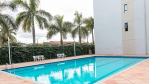 A rectangular swimming pool with a tiled deck, surrounded by palm trees and a white building. - Maria Quitéria Hotel e Flat (Feira de Santana)