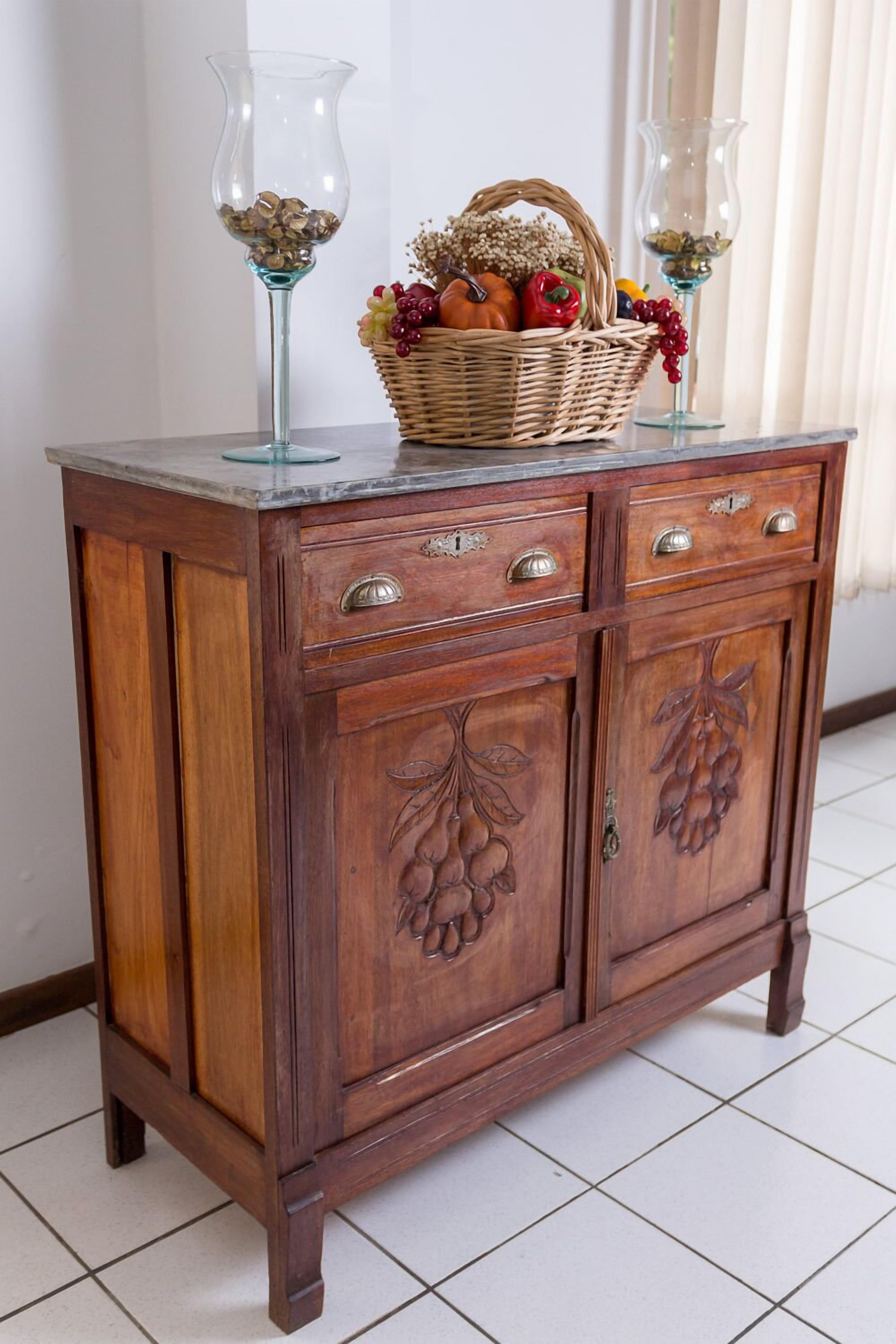 Un meuble en bois doté de deux tiroirs et de deux portes, surmonté d’un vase en verre et d’un panier en osier contenant des fruits et des raisins.