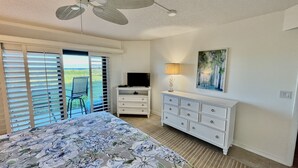 Une chambre à coucher comprenant un lit, une commode, un téléviseur, un ventilateur de plafond et une fenêtre avec des stores.