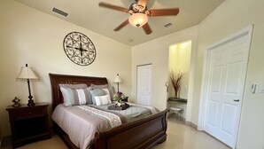 A bedroom with a wooden bed, two bedside tables with lamps, a ceiling fan, a decorative wall piece, and a door leading to another room.