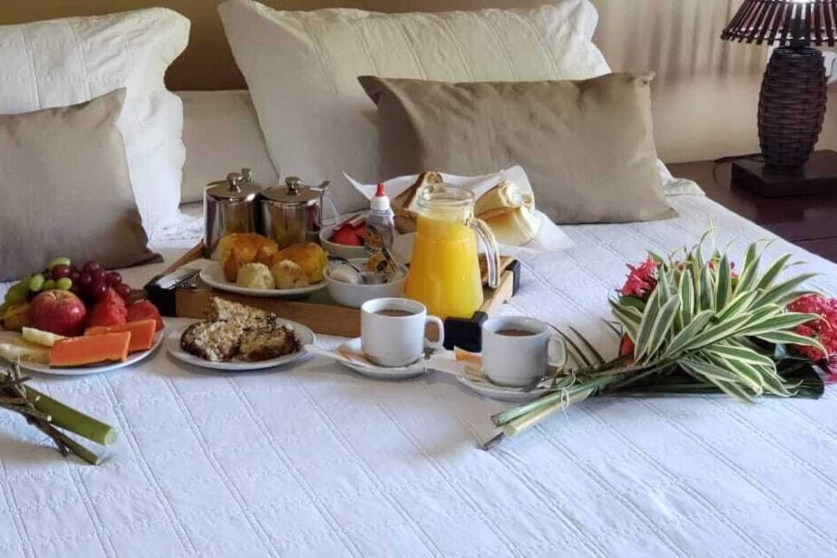 A neatly arranged breakfast table with fruits, pastries, and beverages, set on a bed with pillows and a lamp.