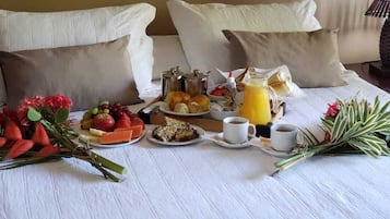Una mesa de desayuno ordenada con frutas, pasteles, café y jugo sobre un mantel blanco en una habitación de hotel.