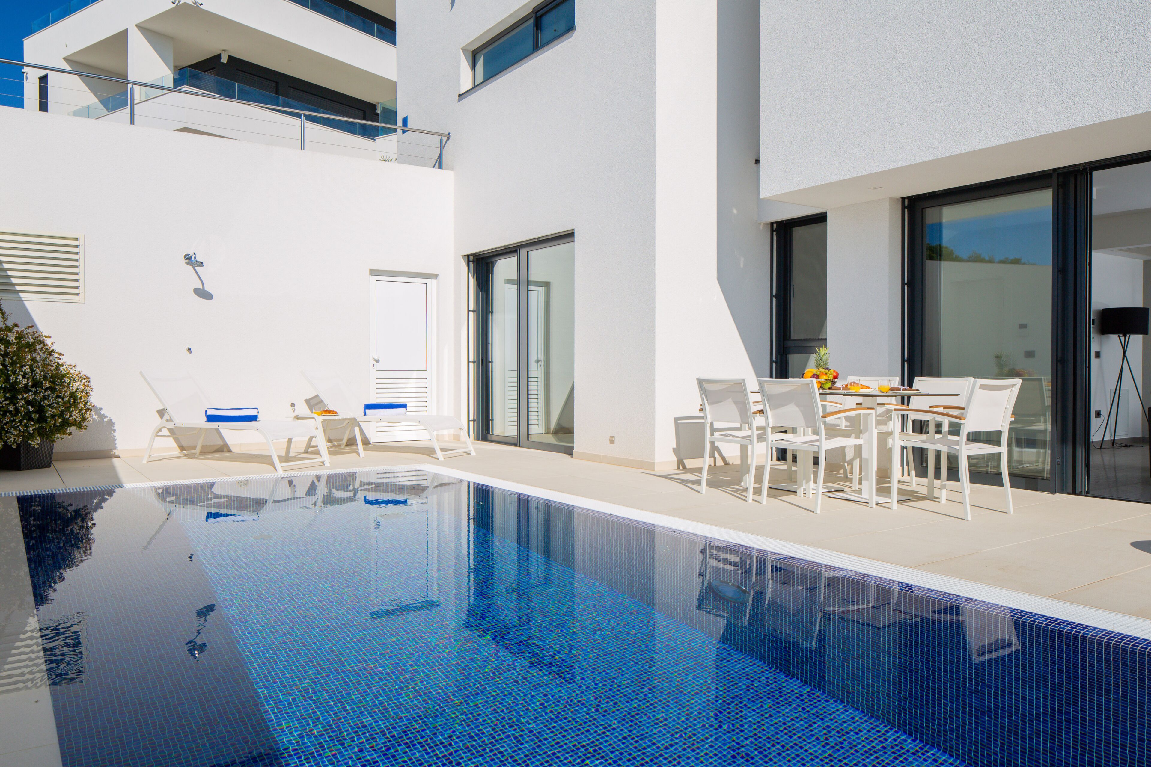 A modern outdoor pool area with lounge chairs and a table, surrounded by a white building with large glass doors.