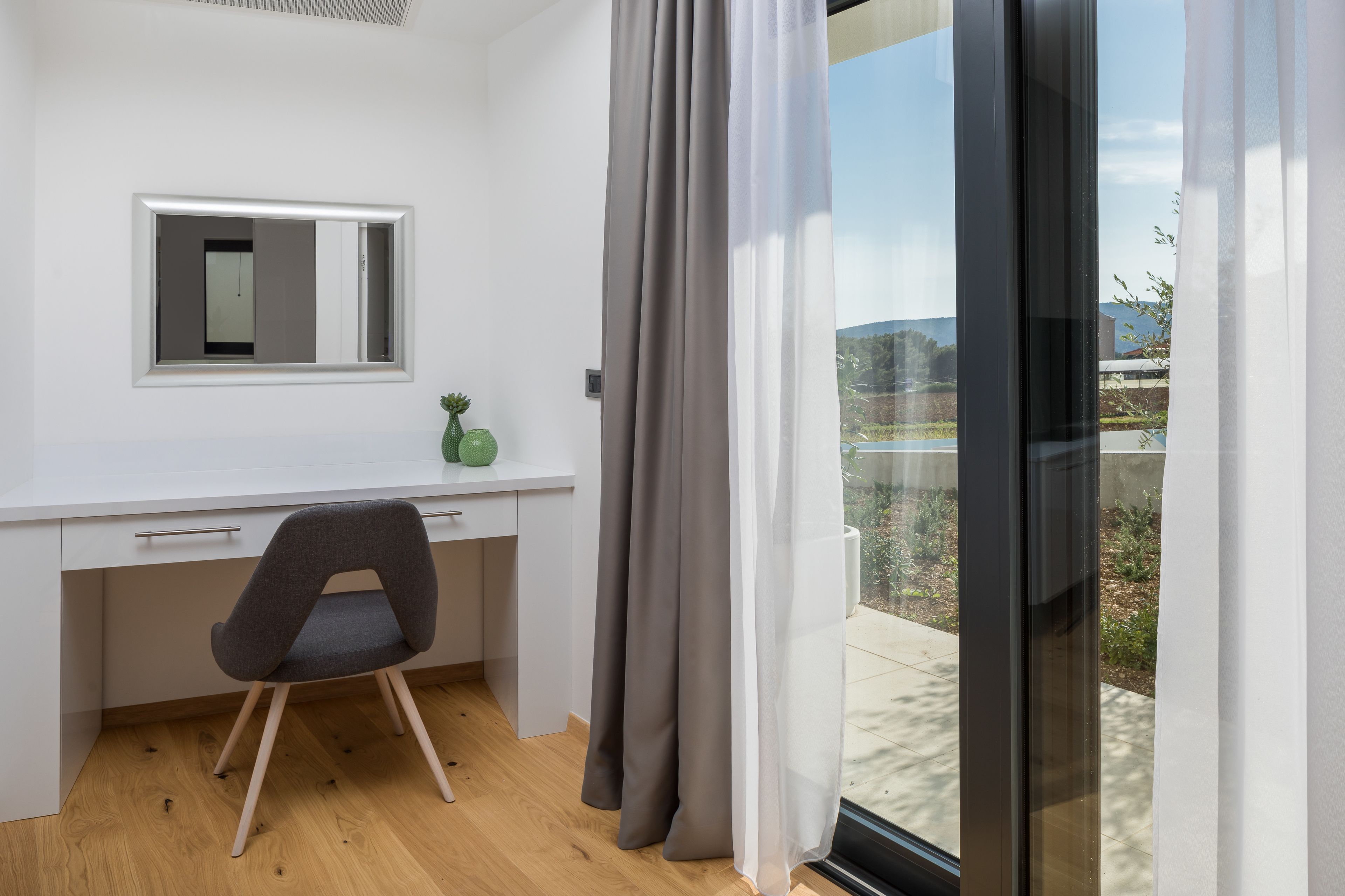 A modern room with a white desk, a grey chair, a small plant, and a large window with curtains.