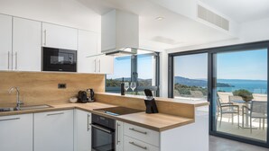 A modern kitchen with white cabinetry, a built-in oven, a microwave, and a large window overlooking a scenic view. - Villas Baotic (Seget)