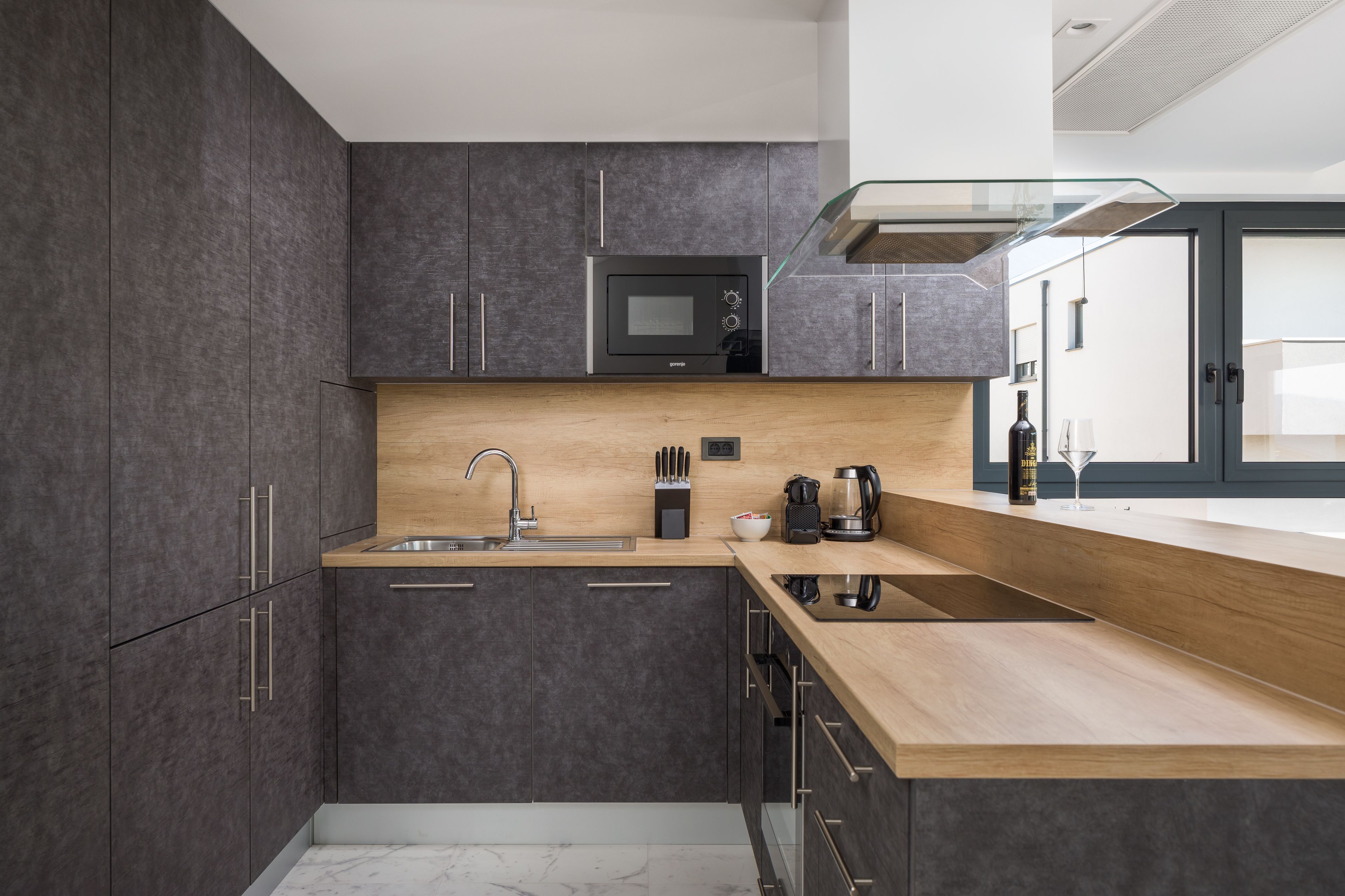 A modern kitchen with dark cabinets, a wooden countertop, a built-in microwave, and a glass-fronted range hood.