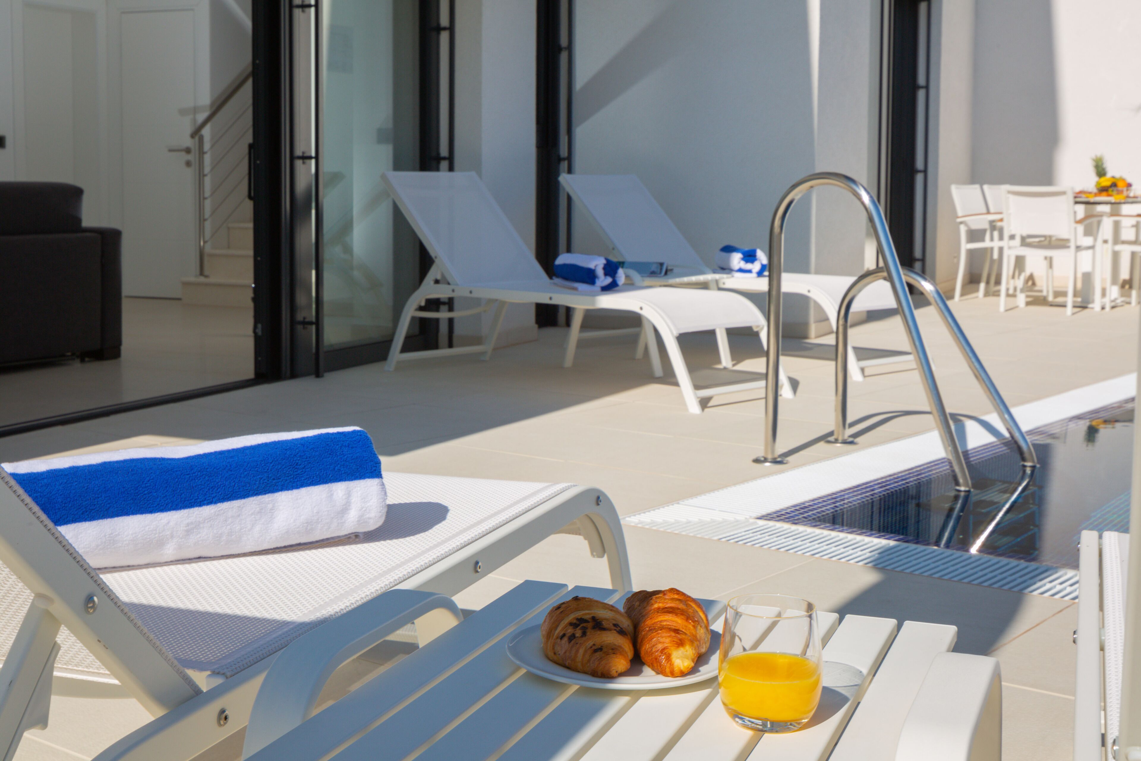 A poolside area with lounge chairs, a towel, croissants, and a glass of orange juice.