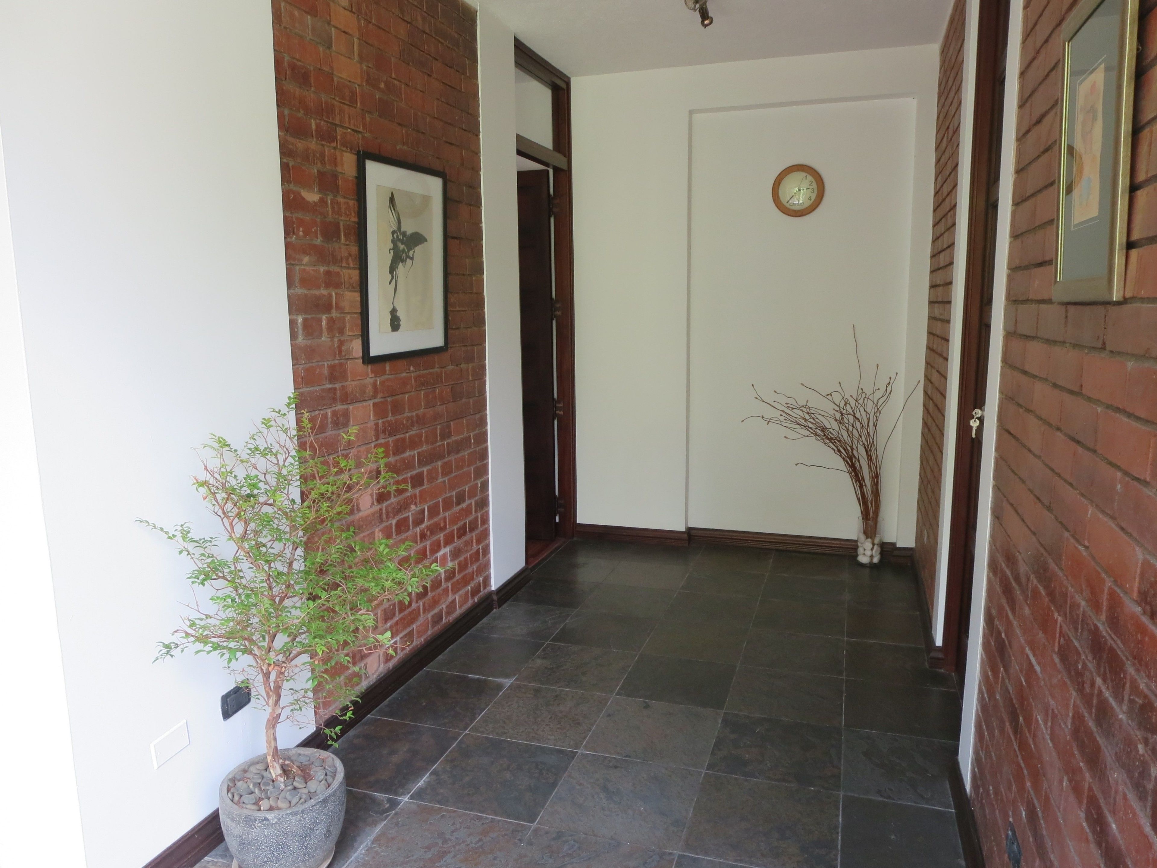 A hallway with a brick wall, a framed picture, a potted plant, a door with a clock, and a tiled floor.