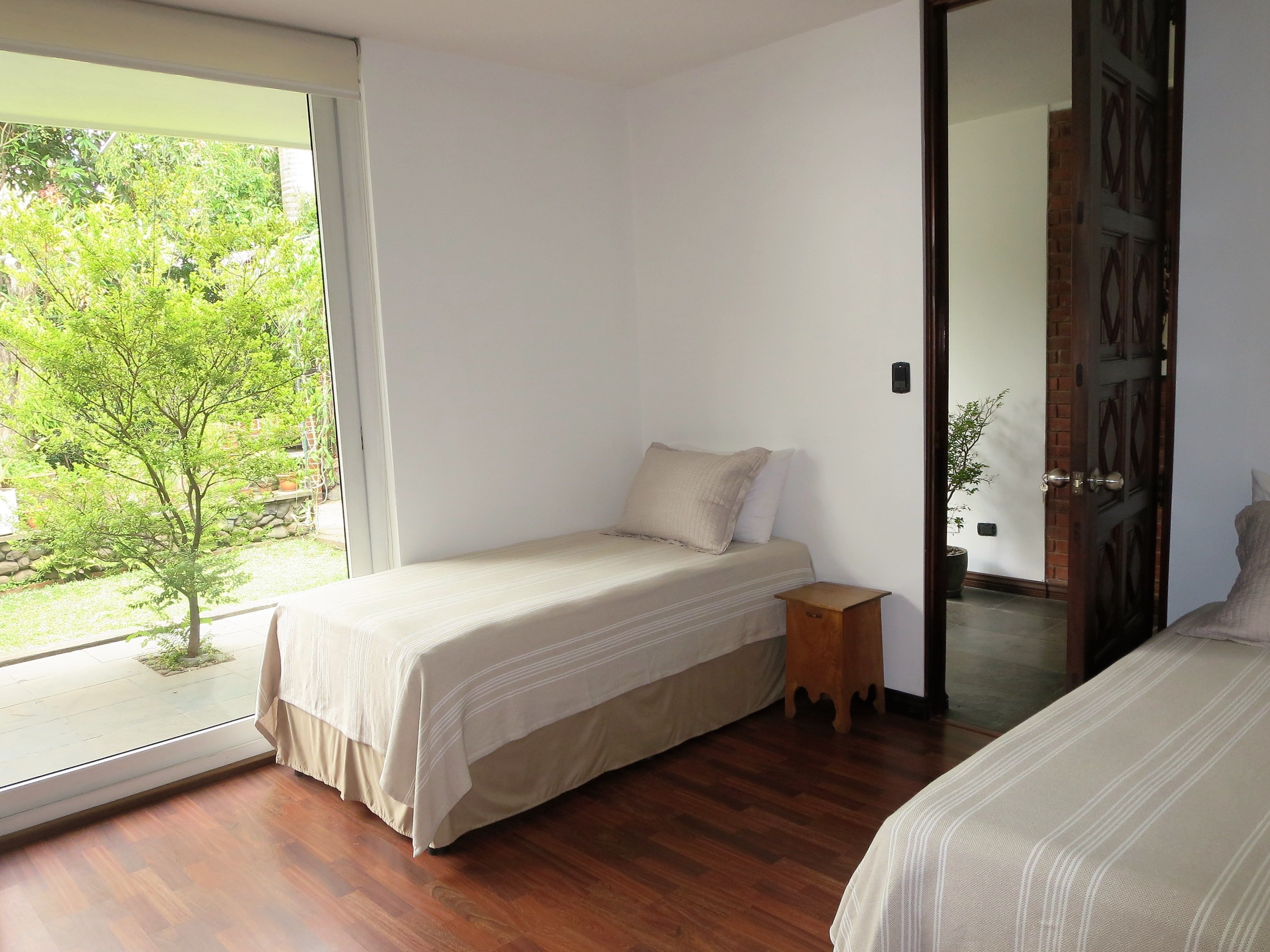 A bedroom with a single bed, a small bedside table, and a large window offering a view of greenery outside.