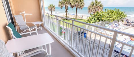 A balcony with a view of the beach, white furniture, and a palm tree.