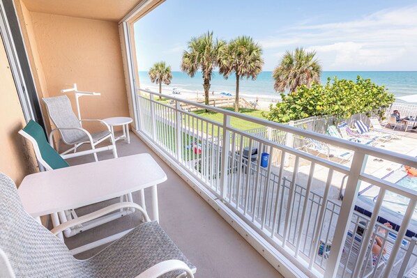 A balcony with a view of the beach, white furniture, and a palm tree.
