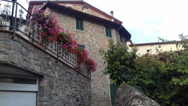 Un edificio de piedra con un balcón decorado con flores rosadas, una puerta de garaje y un árbol de hojas verdes.